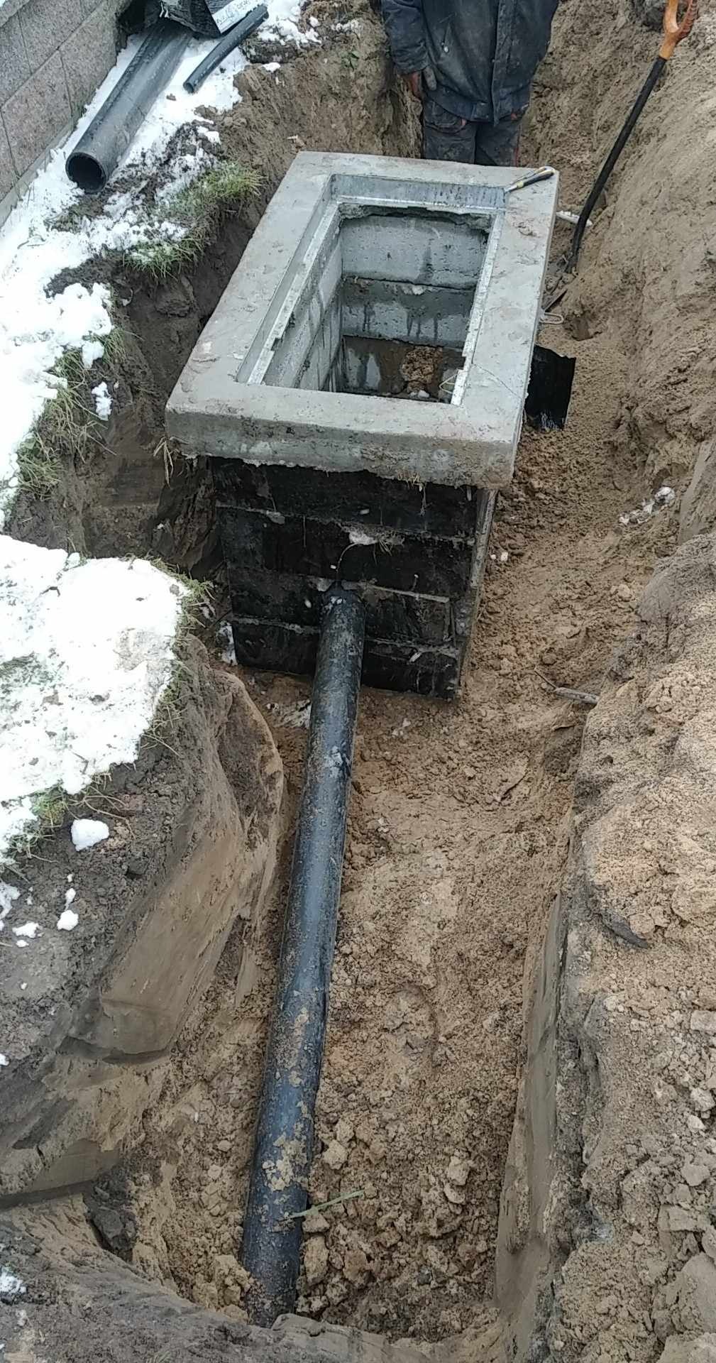 Studzienka kanalizacyjna z rurą w wykopie ziemnym, częściowo ośnieżonym. Widoczna betonowa konstrukcja i czarna rura wchodząca w ziemię. Prace ziemne w toku.