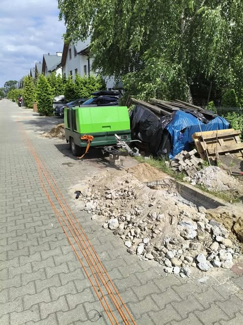 Odkrywka w chodniku z widocznymi kablami i zielonym kompresorem na przyczepie w tle, sugerująca prace instalacyjne na zewnątrz budynku.