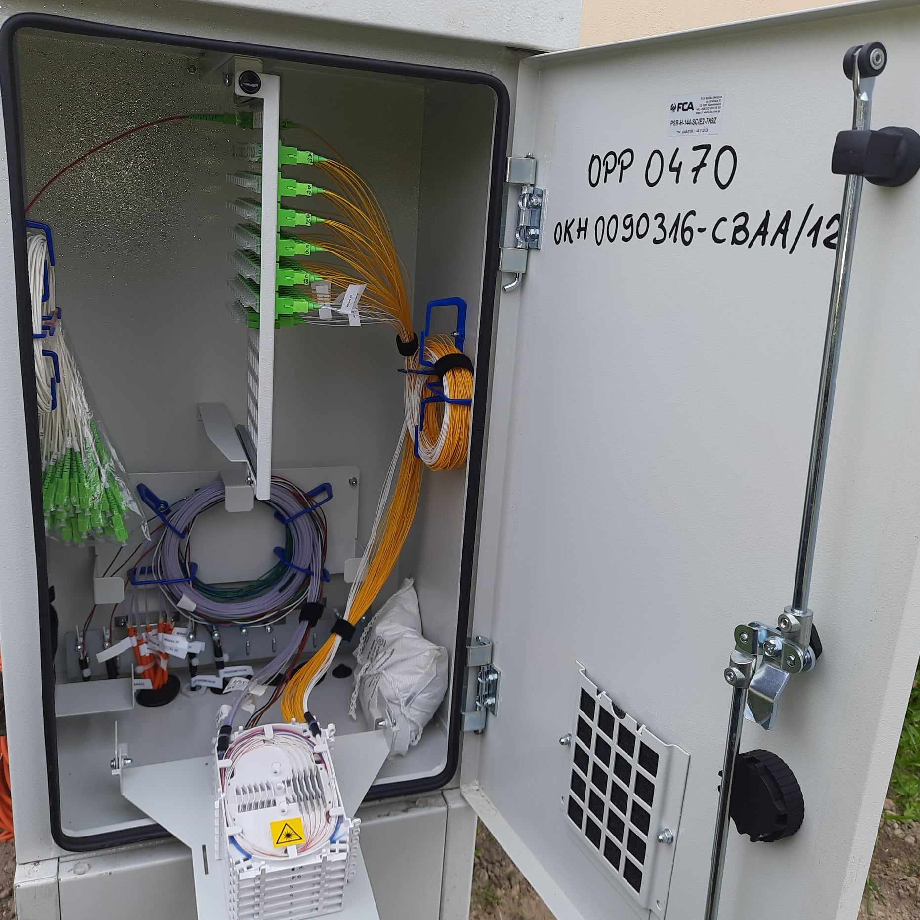 Otwarta skrzynka telekomunikacyjna z licznymi kablami światłowodowymi zakończonymi zielonymi złączkami, uporządkowanymi wewnątrz obudowy. Widoczne oznaczenia na drzwiach.