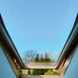 Otwarty okno dachowe z drewnianą ramą, widok na błękitne niebo i drzewa iglaste na zewnątrz.