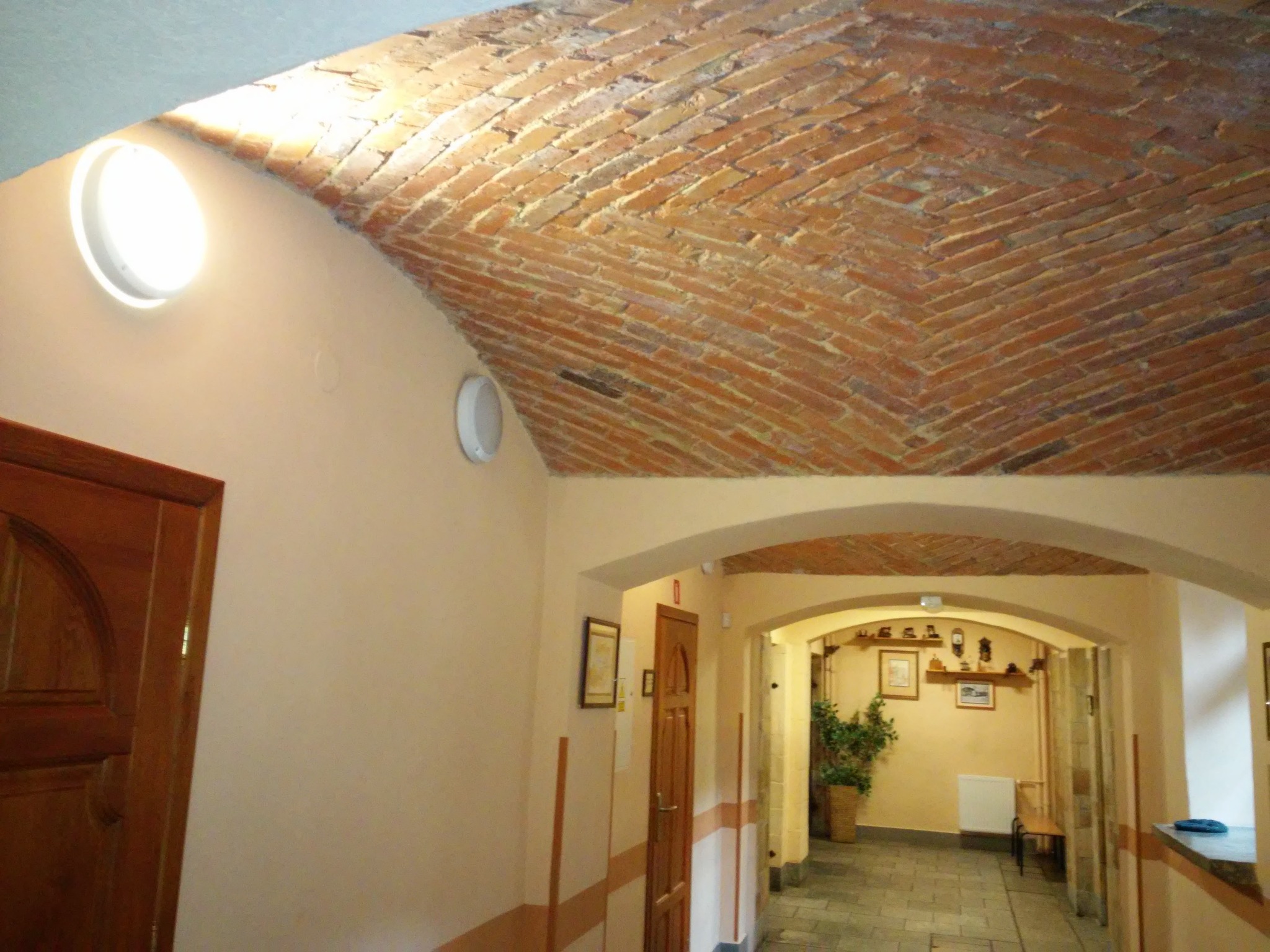 Korytarz z łukowym sufitem wyłożonym cegłą w jodełkę, oświetlony okrągłymi lampami sufitowymi, z widocznymi drewnianymi drzwiami i przejściem do dalszej części budynku.