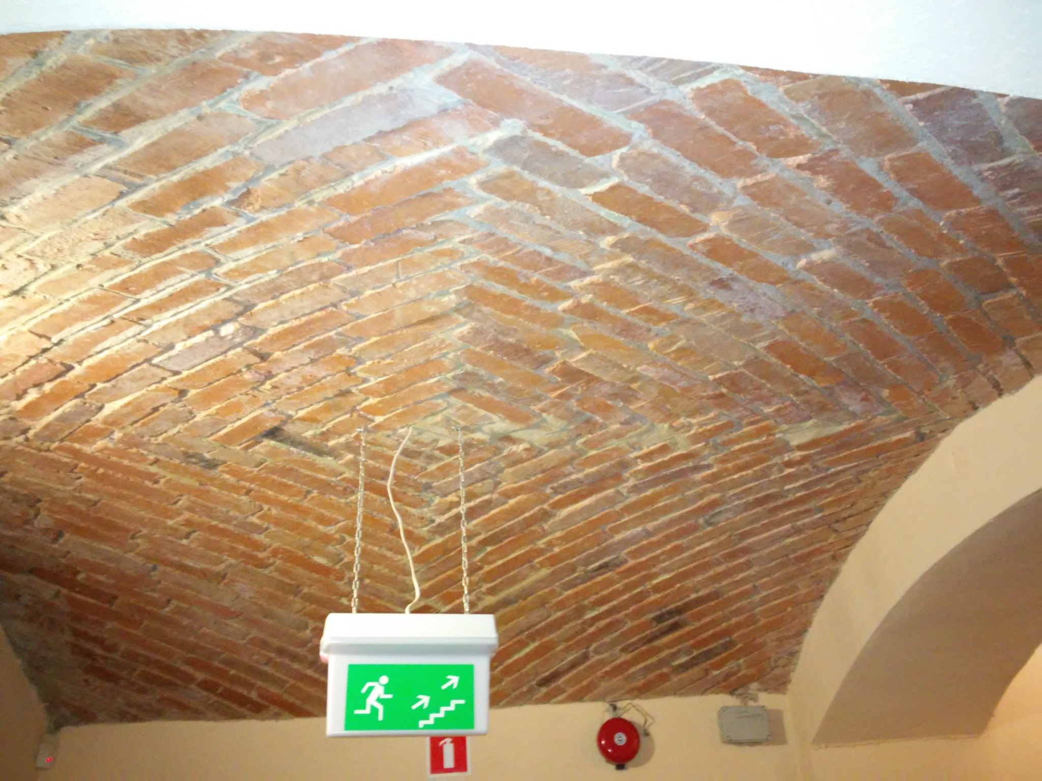 Ceglany, łukowy sufit z zamontowanym podświetlanym znakiem ewakuacyjnym na łańcuszku, obok czerwony alarm pożarowy.