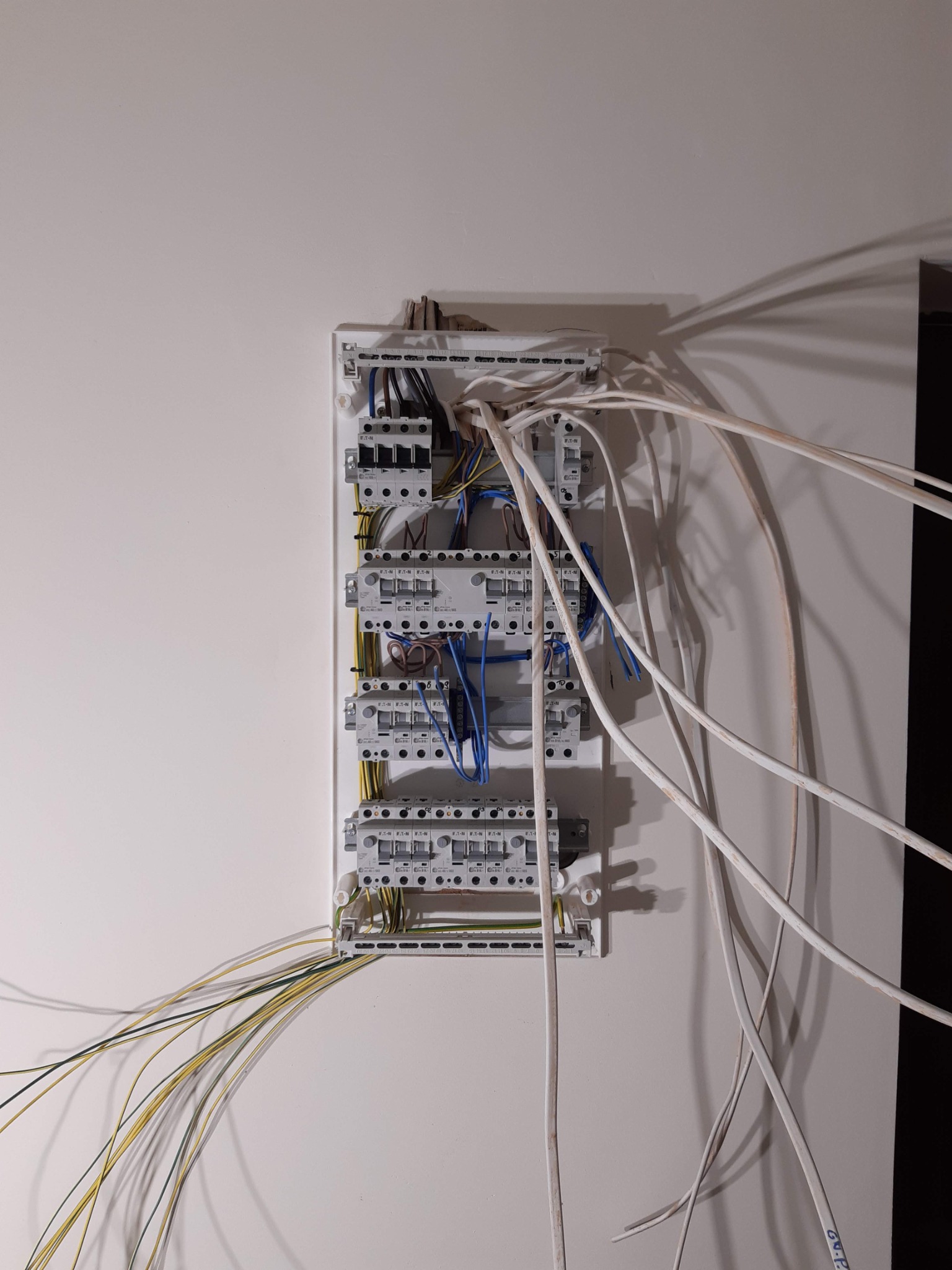 Rozdzielnica elektryczna z wystającymi przewodami na białej ścianie, widoczne zabezpieczenia Eaton i przewody w kolorach żółto-zielonym, niebieskim i białym.