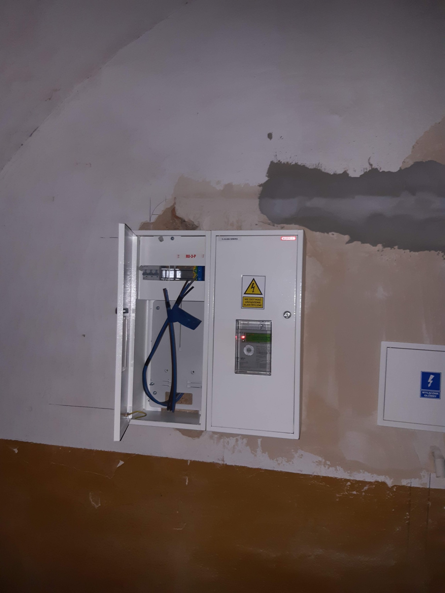 Otwarta skrzynka rozdzielcza z widocznymi przewodami i bezpiecznikami, zamontowana na odrapanej ścianie w trakcie remontu, obok dodatkowa skrzynka z napisem 'Wyłącznik główny'.