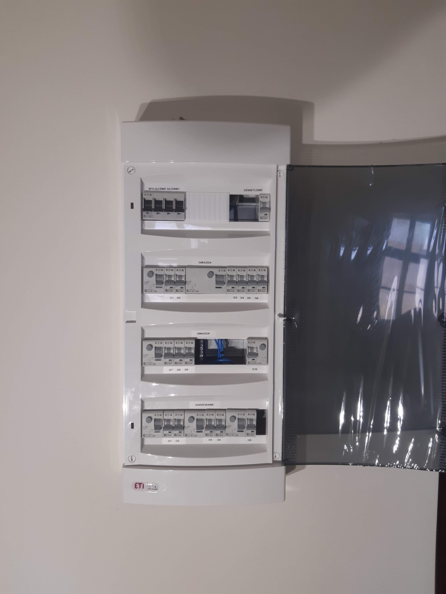 Rozdzielnica elektryczna z widocznymi bezpiecznikami i wyłącznikami oznaczonymi kategoriami obwodów: wyłącznik główny, oświetlenie, gniazda, ogrzewanie; częściowo otwarta osłona ochronna.