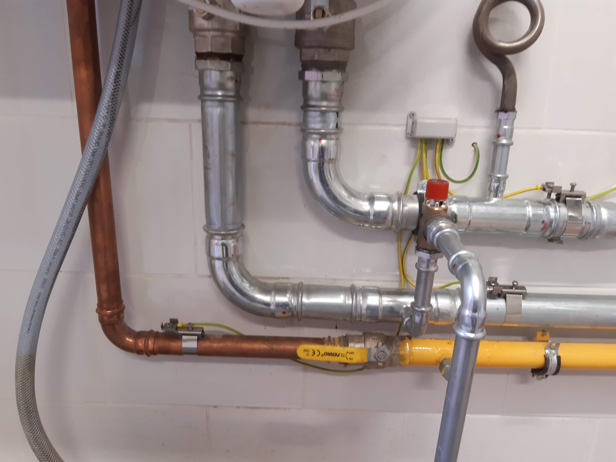 Złożony system rur z różnych materiałów (miedź, stal, plastik) z zaworami, złączkami i przewodami uziemiającymi na tle białych kafelków.