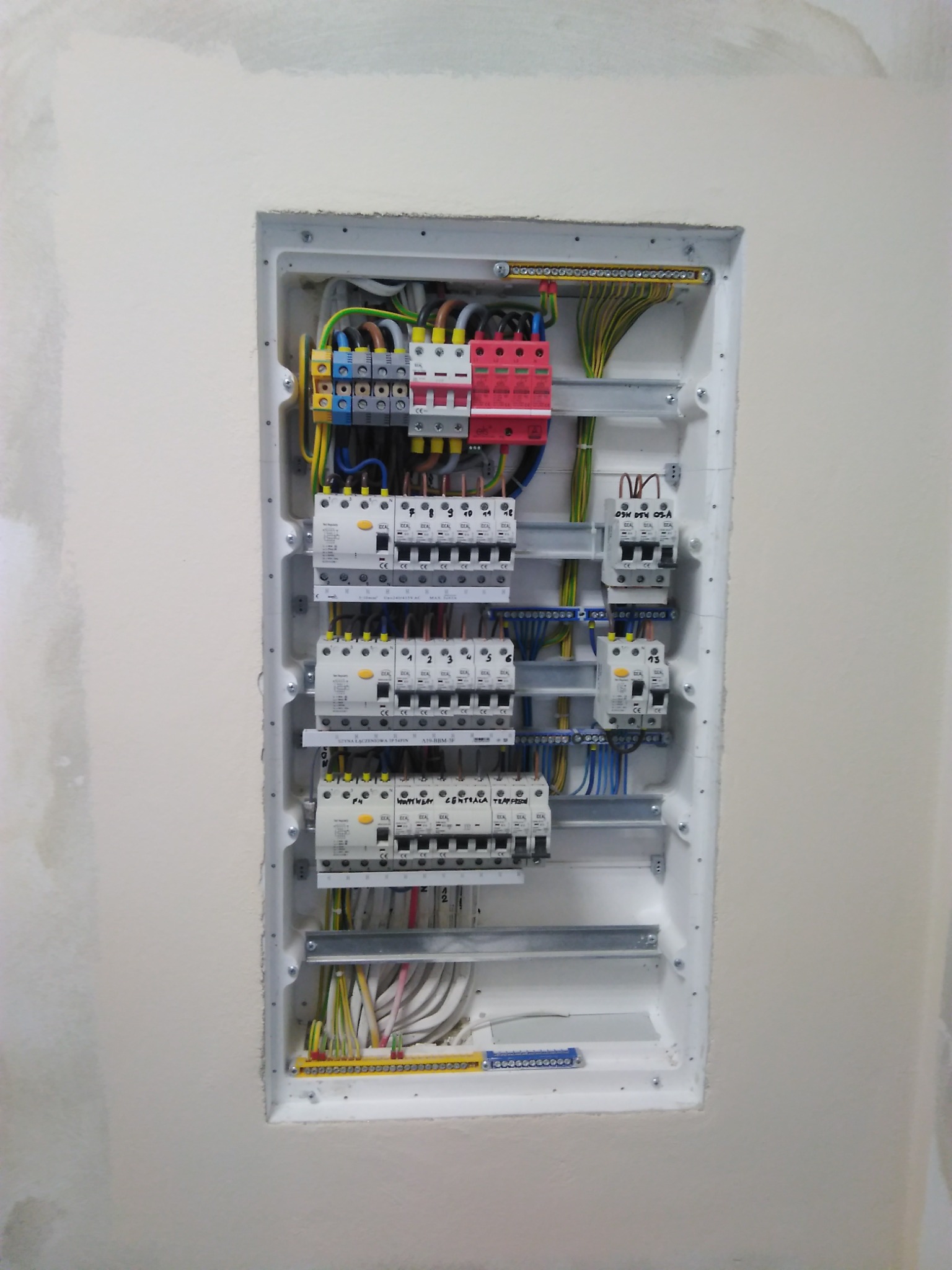 Wnętrze skrzynki rozdzielczej w ścianie, widoczne bezpieczniki, wyłączniki, przewody i szyny montażowe, instalacja w trakcie podłączenia.