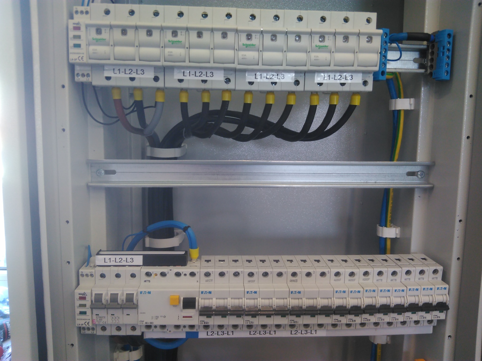 Otwarta skrzynka rozdzielcza z bezpiecznikami, widoczne przewody z żółtymi końcówkami, szyna montażowa i aparatura modułowa marek Schneider i Eaton.