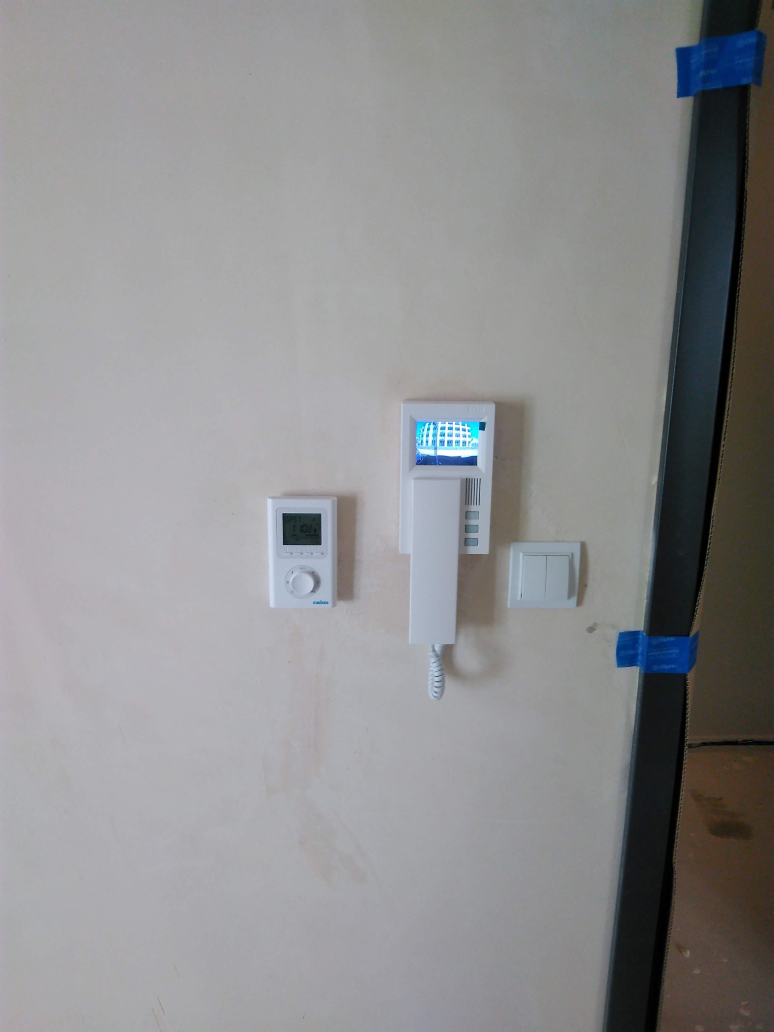 Biały panel domofonu z wyświetlaczem, termostat pokojowy i włącznik światła zamontowane na ścianie w trakcie remontu, zabezpieczone niebieską taśmą malarską