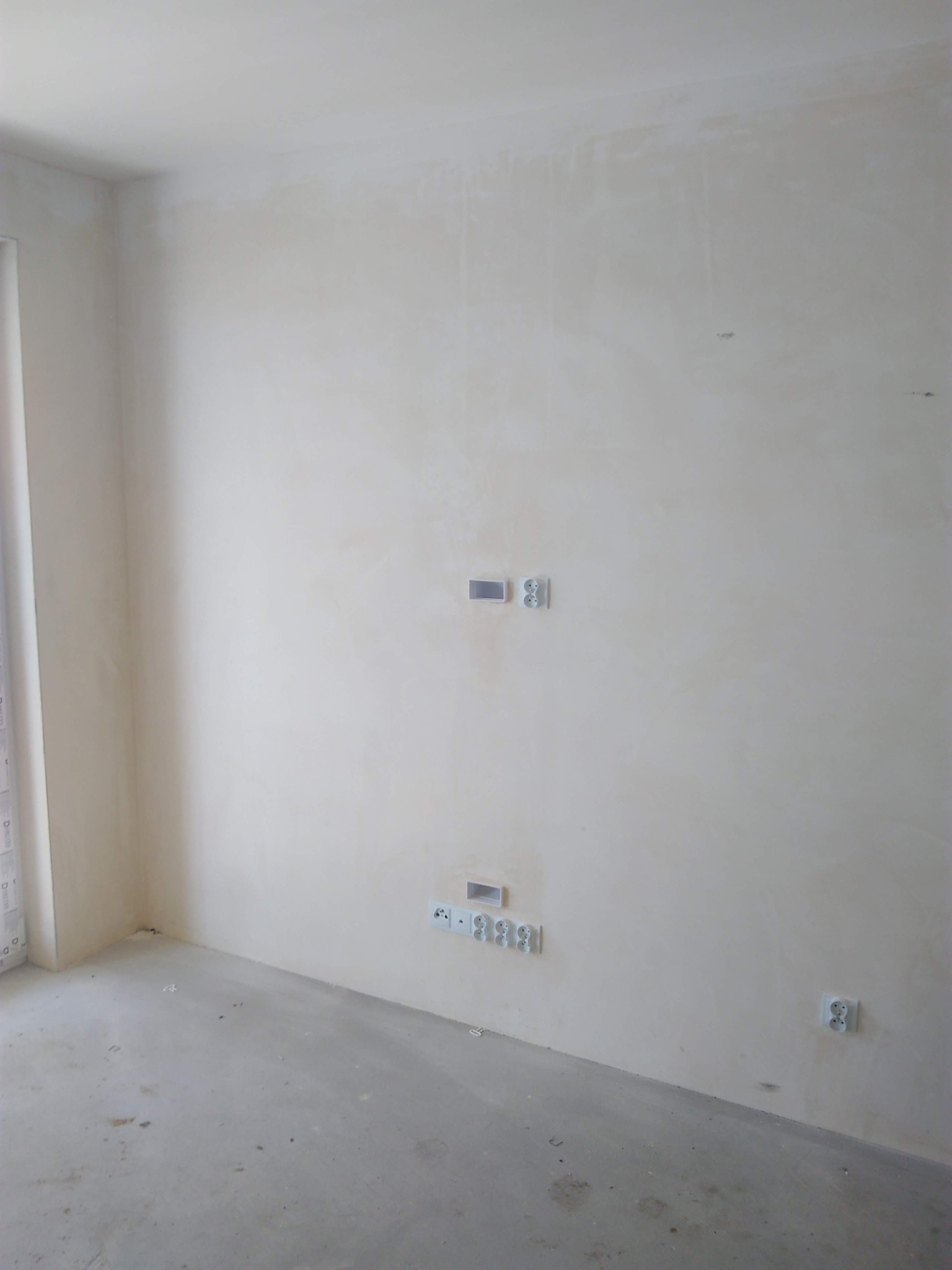 Świeżo otynkowane wnętrze z zamontowanymi gniazdkami elektrycznymi i puszką instalacyjną, widoczne ślady po pracach budowlanych na podłodze.