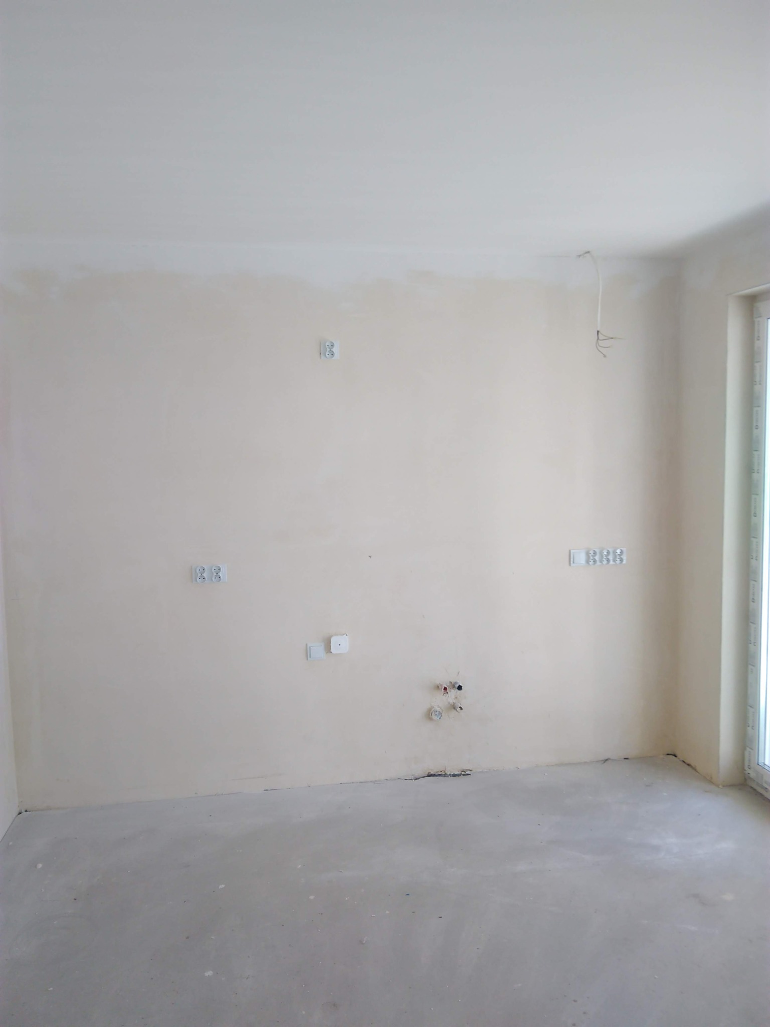 Surowe wnętrze pokoju z widocznymi puszkami elektrycznymi, gniazdkami i wyprowadzonymi przewodami, ściany przygotowane pod malowanie, surowa podłoga, okno balkonowe z prawej strony.