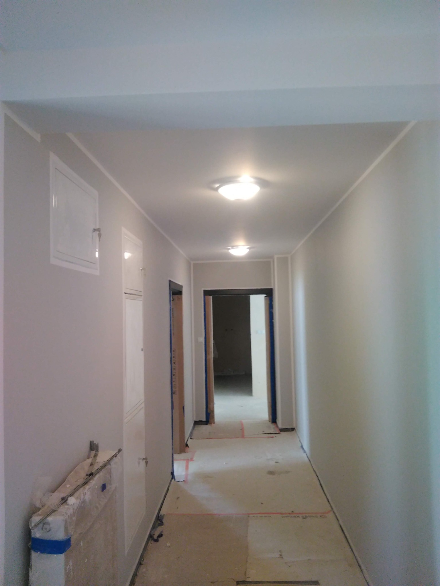 Korytarz w trakcie remontu, widoczne nowe oświetlenie sufitowe, zabudowane skrzynki instalacyjne na ścianie, zabezpieczone ościeżnice drzwiowe i fragment podłogi z resztkami po pracach.