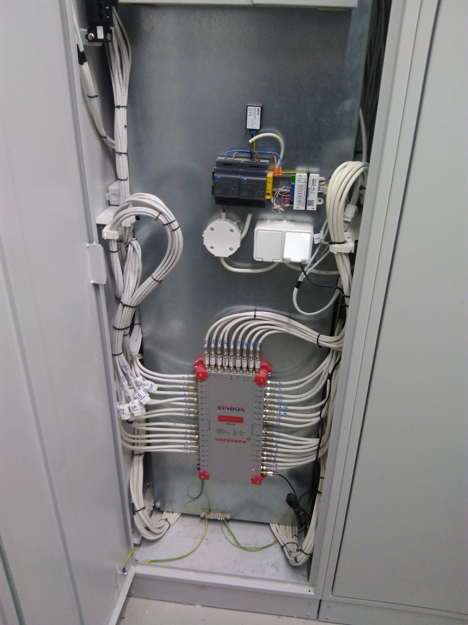 Otwarta szafa elektryczna z zamontowanym rozdzielaczem kablowym LindoX, licznymi przewodami oznaczonymi numerami, zabezpieczeniami i elementami instalacji elektrycznej na metalowej płycie.