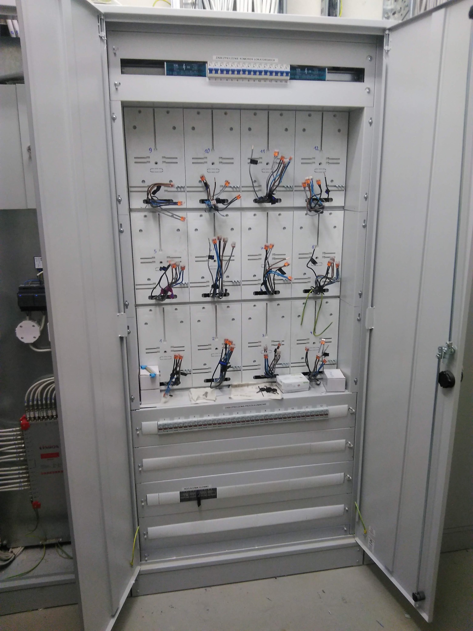 Otwarta szafa elektryczna z widocznymi zabezpieczeniami, przewodami i złączkami, częściowo podłączonymi do ponumerowanych modułów, z widoczną listwą zaciskową u góry i elementami instalacji po...