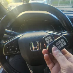 Dwa kluczyki samochodowe Honda trzymane w dłoni na tle kierownicy z logo marki, widoczne elementy deski rozdzielczej i szyba przednia z odbiciami światła.