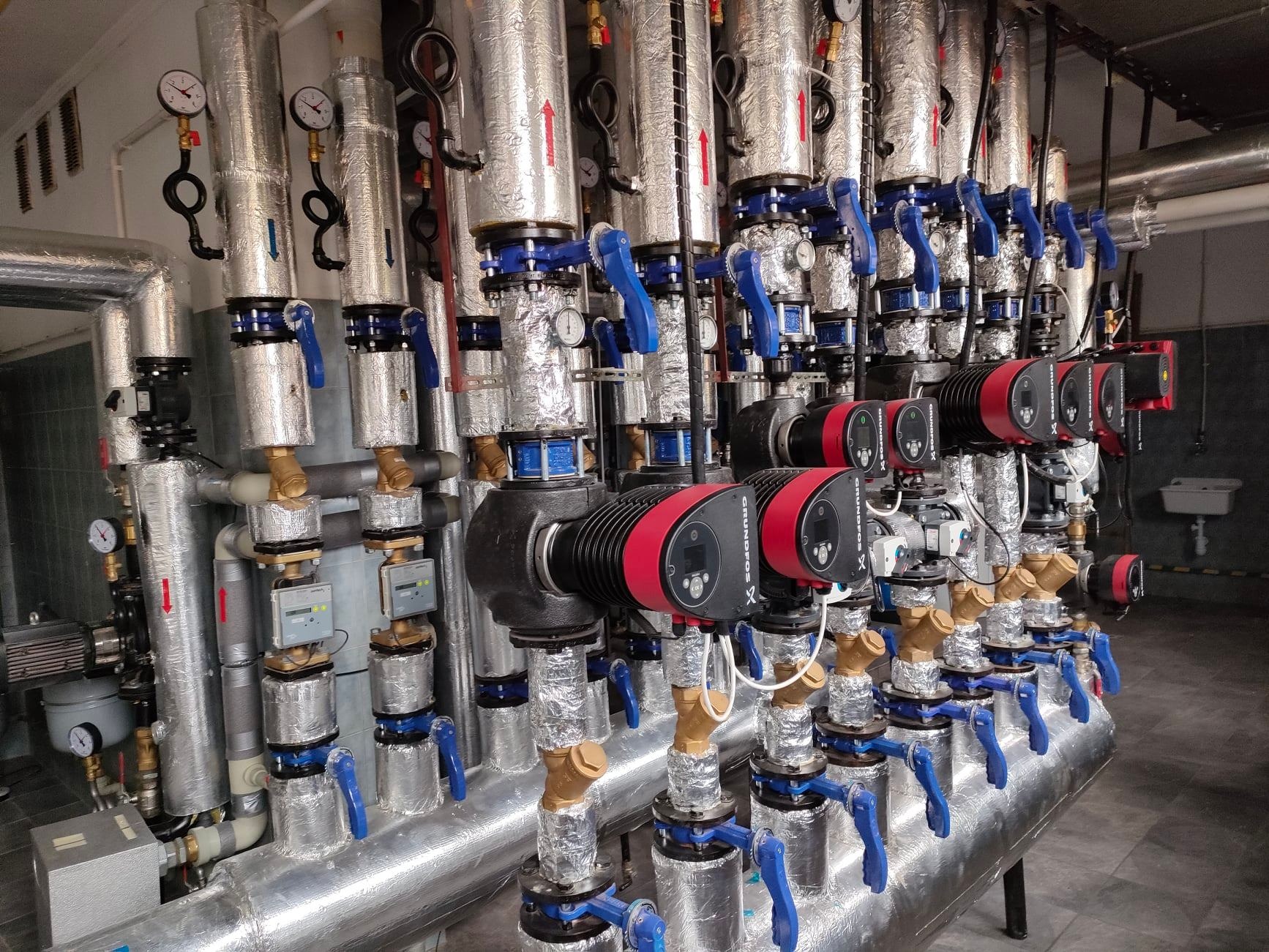 Rozbudowana instalacja grzewcza z licznymi pompami obiegowymi Grundfos, zaworami z niebieskimi uchwytami i izolowanymi rurami, wyposażona w manometry i liczniki ciepła, całość w pomieszczeniu...