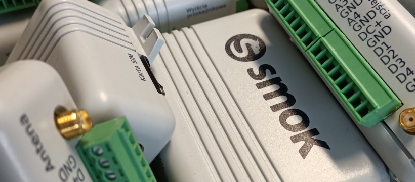 Zbliżenie na moduły sterujące z gniazdami antenowymi, złączami śrubowymi i slotem karty SIM, oznaczone logo 'Smok'.