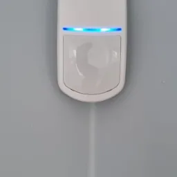 Biały czujnik alarmowy z niebieskim paskiem LED zamontowany w rogu szarego pokoju.