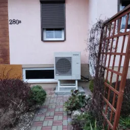 Zewnętrzna jednostka klimatyzacji Samsung zamontowana przy różowej ścianie budynku z brązowymi roletami i numerem 280b, widoczna z brukowanego chodnika i ogrodu.