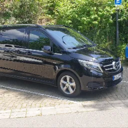Czarny van Mercedes V-klasy zaparkowany na brukowanym parkingu, widoczny znak parkingu, obok stoi niebieski samochód osobowy.