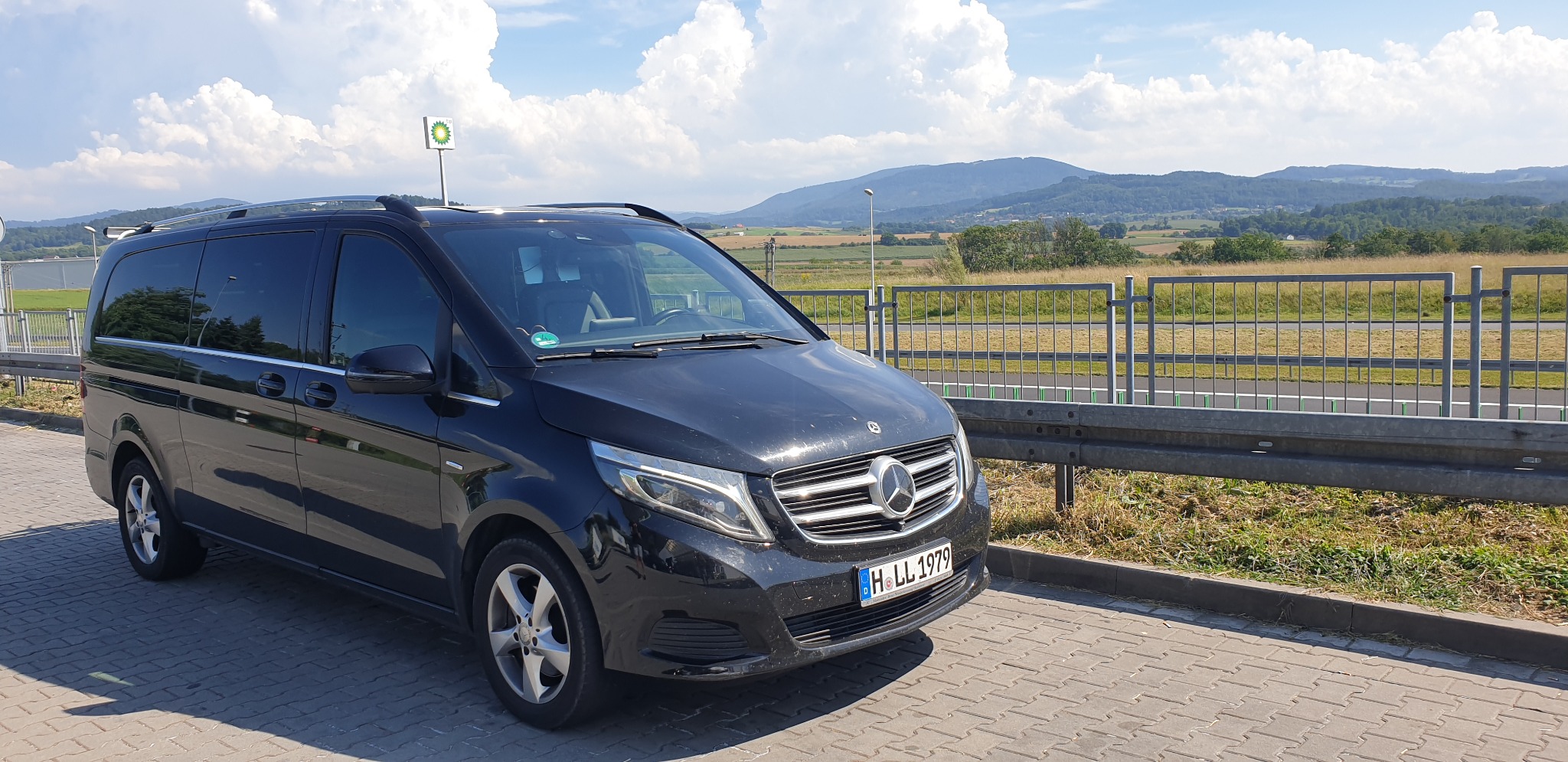 Czarny Mercedes V-klasa zaparkowany na kostce brukowej z widokiem na pagórkowaty krajobraz i błękitne niebo z chmurami, widoczny znak BP w tle.