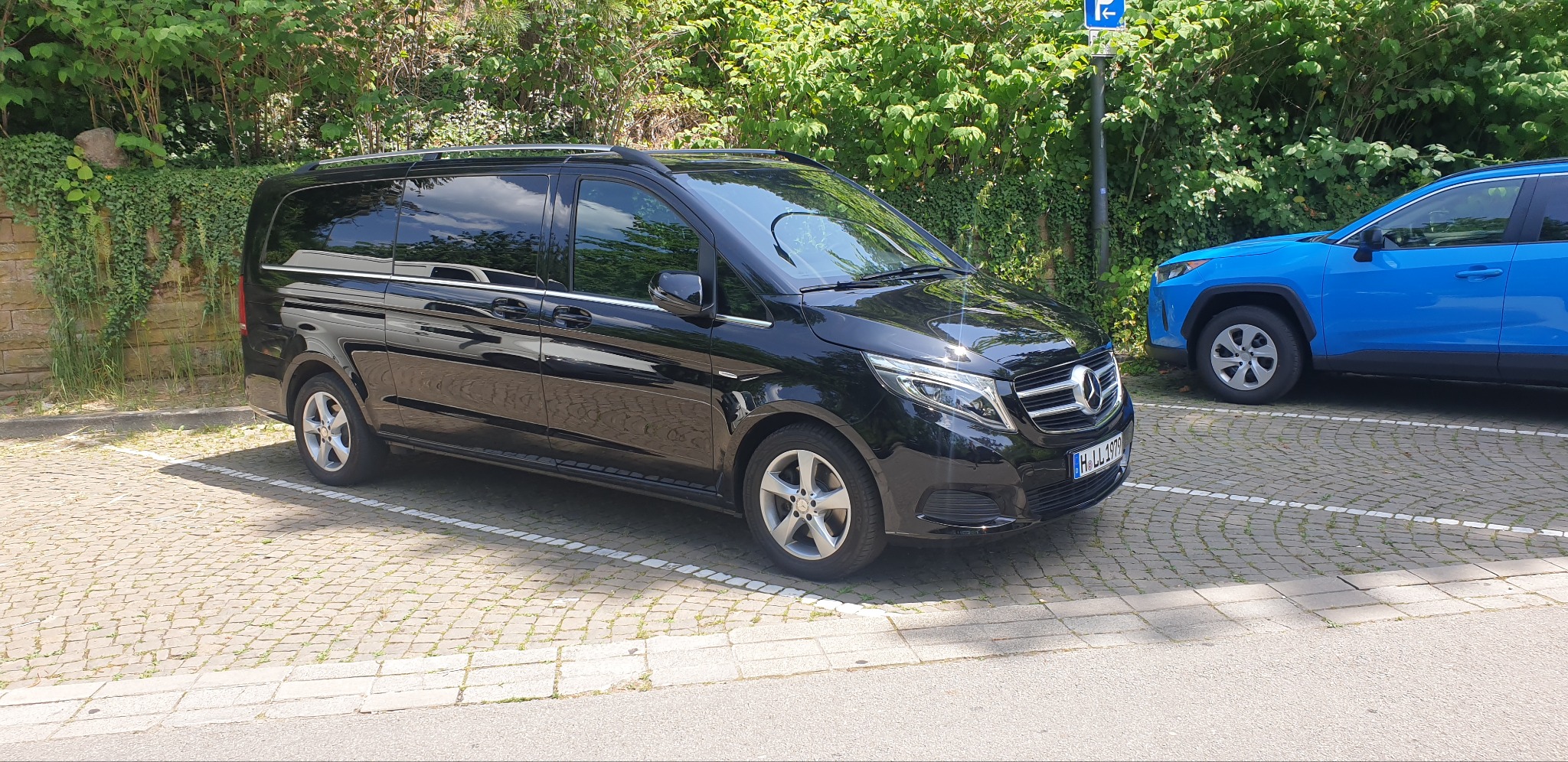 Czarny van Mercedes V-klasy zaparkowany na brukowanym parkingu, widoczny znak parkingu, obok stoi niebieski samochód osobowy.