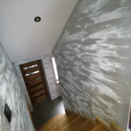 Klatka schodowa z drewnianymi schodami i ścianami wykończonymi dekoracyjną szarą farbą z efektem rozmycia, widoczna drewniana ościeżnica drzwi z przeszkleniami.