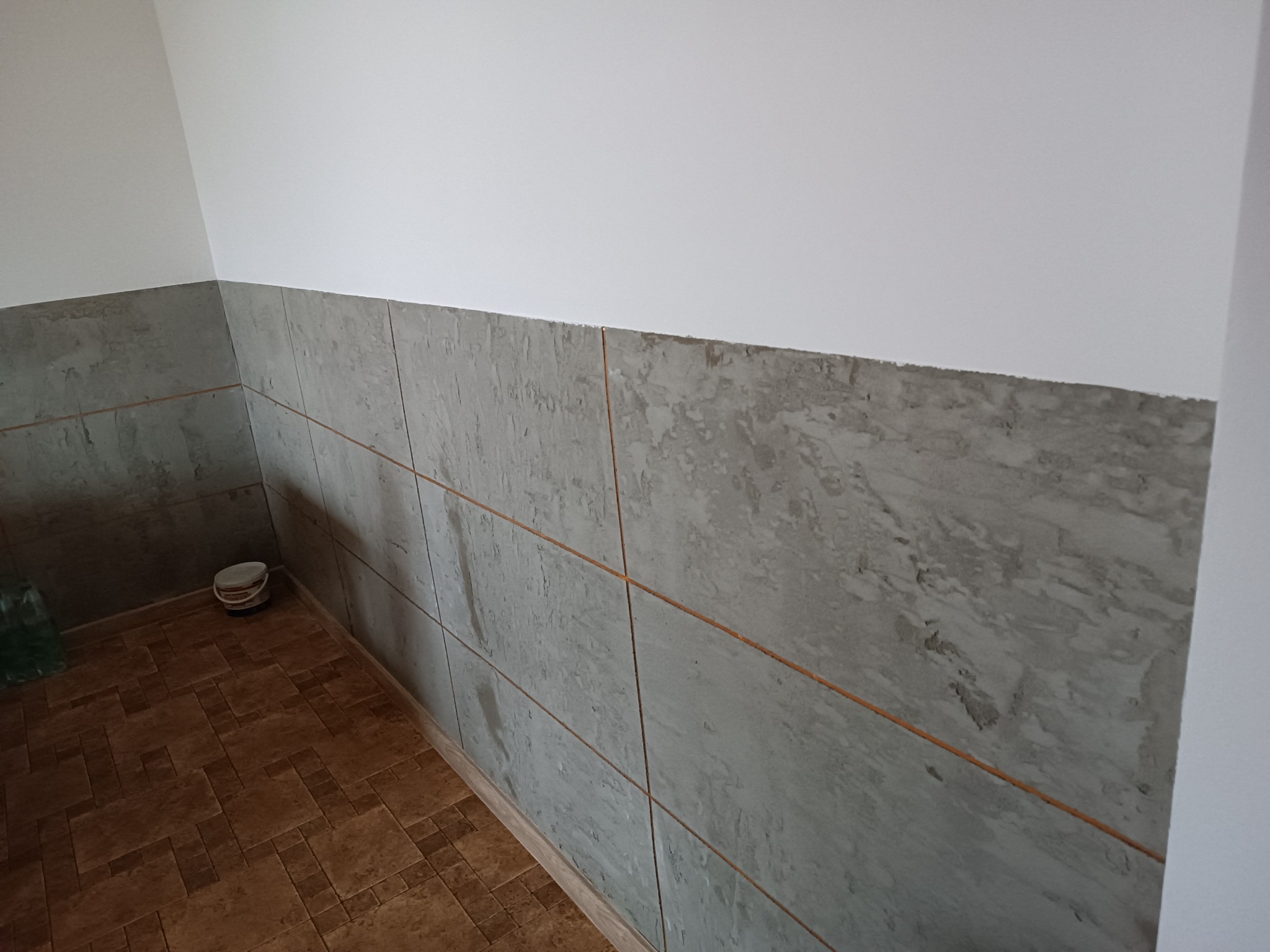 Wykończona dolna część ściany z szarymi płytami imitującymi beton, z widoczną spoiną w kolorze miedzianym, biała ściana powyżej i brązowa podłoga z wzorem płytek, w rogu stoi wiadro.