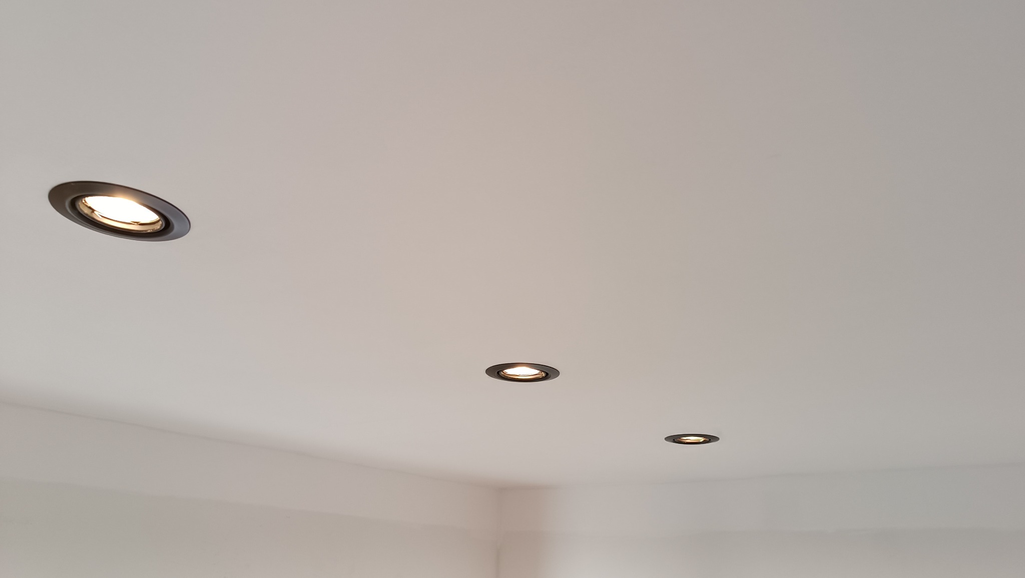 Biały sufit z trzema okrągłymi, wpuszczanymi lampami halogenowymi z czarnymi obwódkami, rozmieszczonymi w strategicznych punktach pomieszczenia.