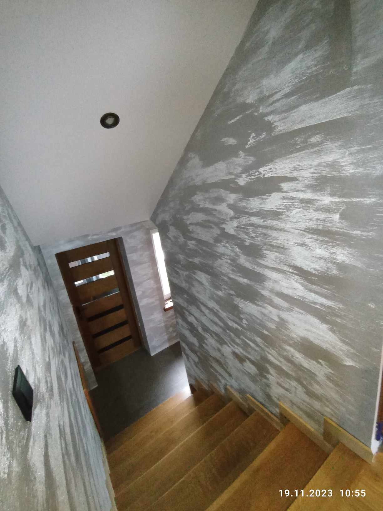 Klatka schodowa z drewnianymi schodami i ścianami wykończonymi dekoracyjną szarą farbą z efektem rozmycia, widoczna drewniana ościeżnica drzwi z przeszkleniami.