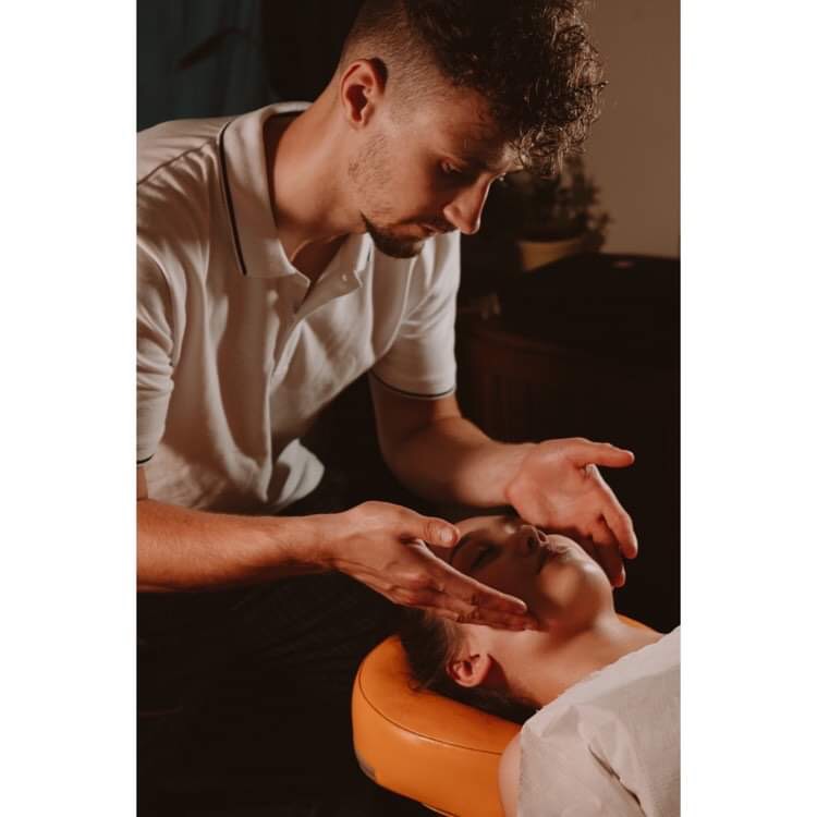 Mężczyzna wykonuje masaż twarzy młodej kobiety leżącej na pomarańczowej poduszce, widoczne dłonie masażysty i fragment białego prześcieradła.