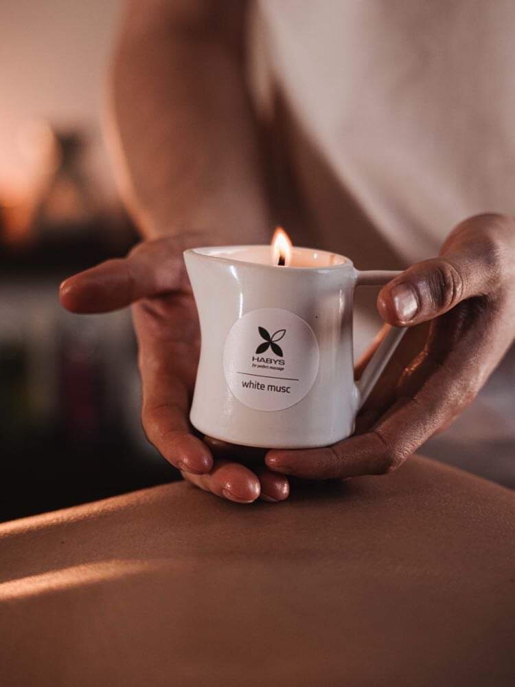 Osoba trzyma ceramiczny dzbanuszek z płonącą świeczką zapachową marki Habys, przygotowując się do zabiegu na plecach klienta.