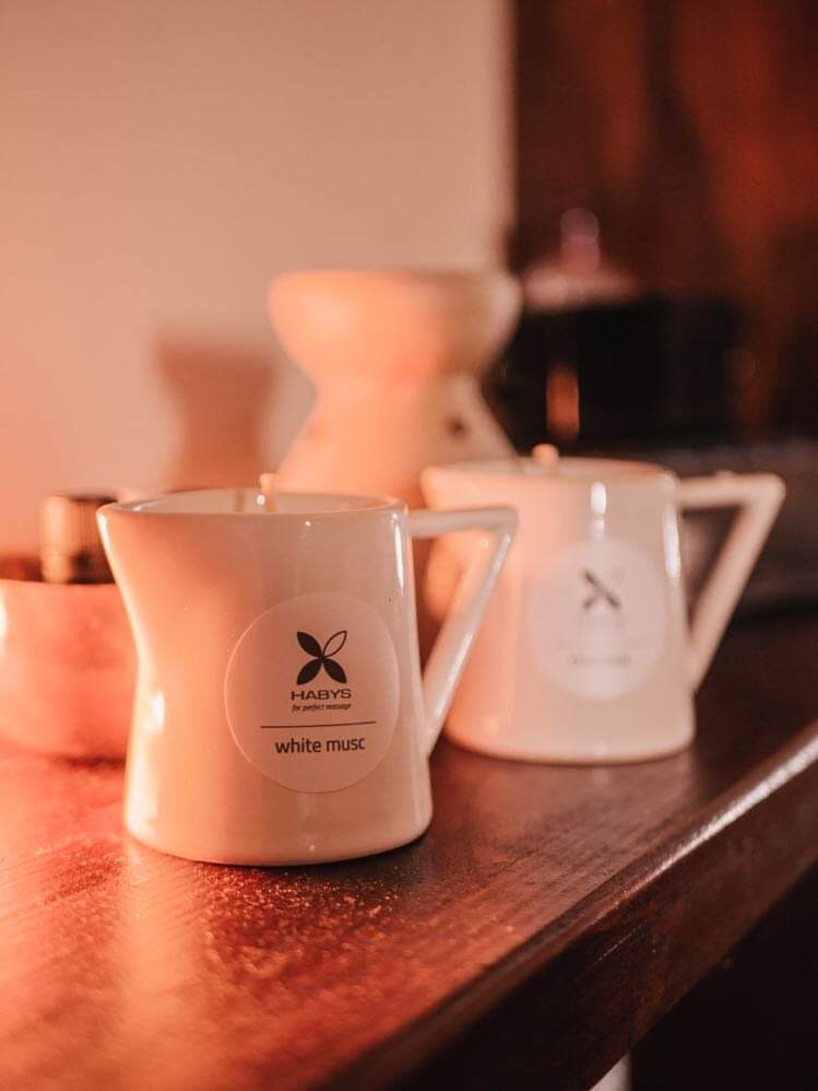 Dwa kremowe dzbanki z etykietami 'Habys white musc' stoją na drewnianej powierzchni, w tle dyfuzor zapachowy i ciemny przedmiot.