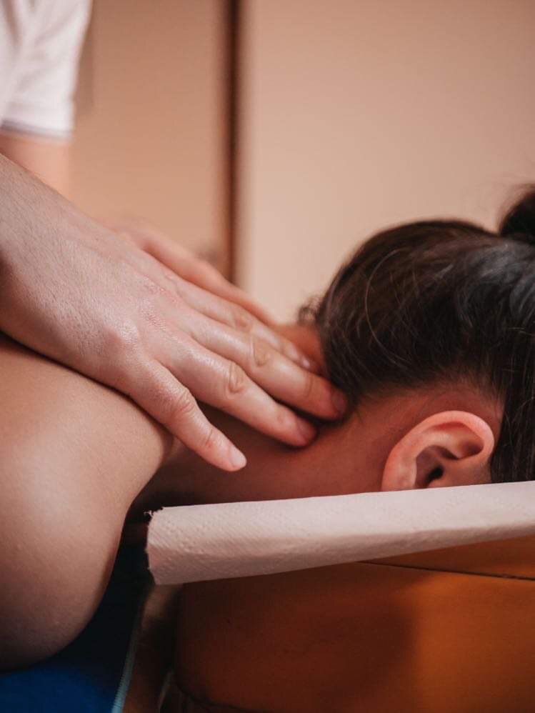 Dłonie terapeuty uciskają szyję pacjentki leżącej na stole z jednorazowym podkładem, widok z boku.