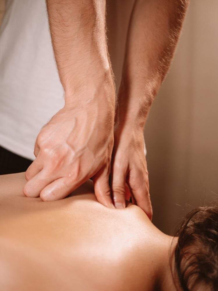 Masażysta uciska kciukami mięśnie pleców klienta; widoczne dłonie i fragment koszulki masażysty, ciepłe światło