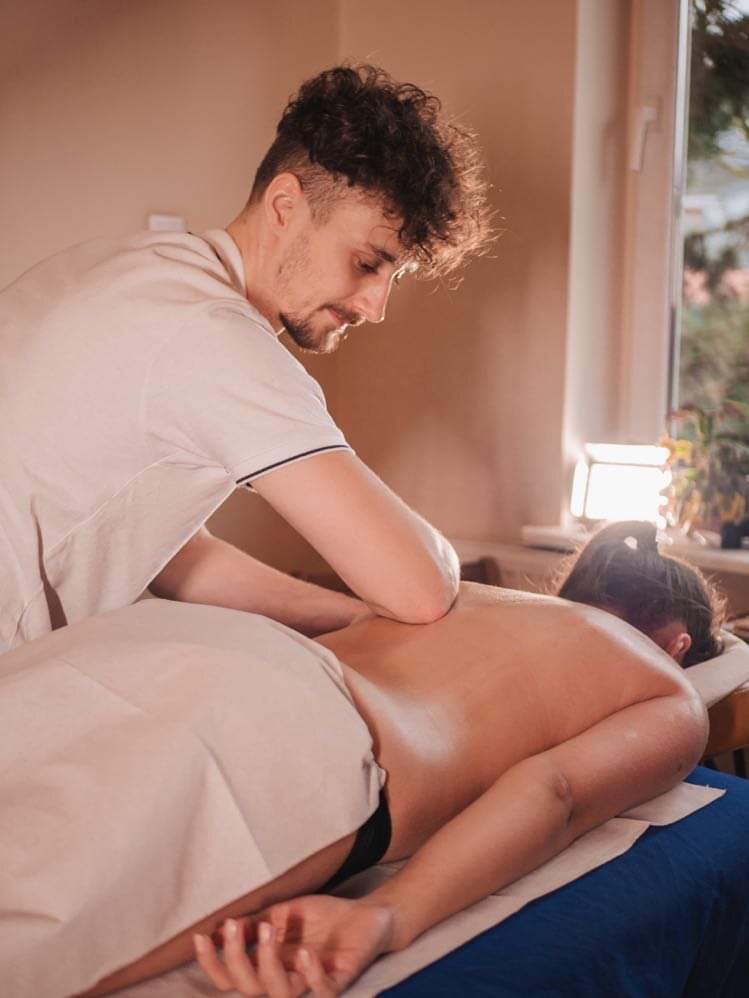 Mężczyzna w białej koszulce wykonuje masaż pleców kobiecie leżącej na niebieskim prześcieradle w gabinecie z oknem w tle.