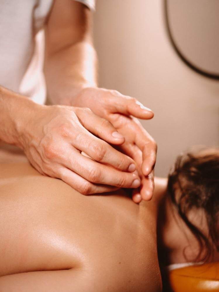 Dłonie terapeuty uciskają plecy pacjentki podczas sesji relaksacyjnej, widoczne włosy pacjentki i fragment okrągłego elementu na ścianie.