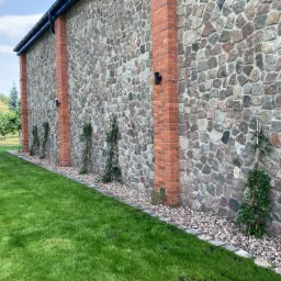 Kamienno-ceglana ściana budynku z oświetleniem zewnętrznym i pnącą roślinnością, obok trawnik i drzewa owocowe.