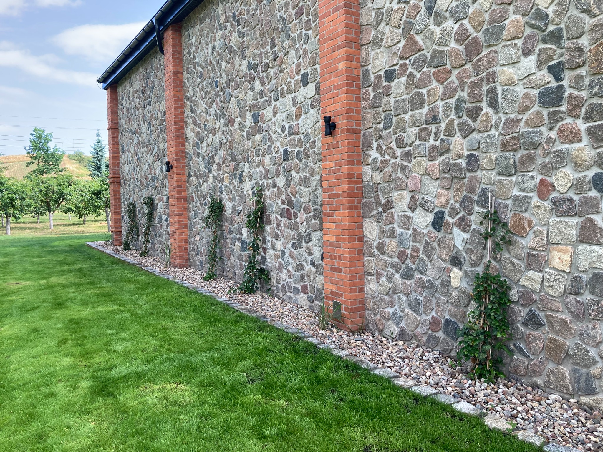 Kamienno-ceglana ściana budynku z oświetleniem zewnętrznym i pnącą roślinnością, obok trawnik i drzewa owocowe.