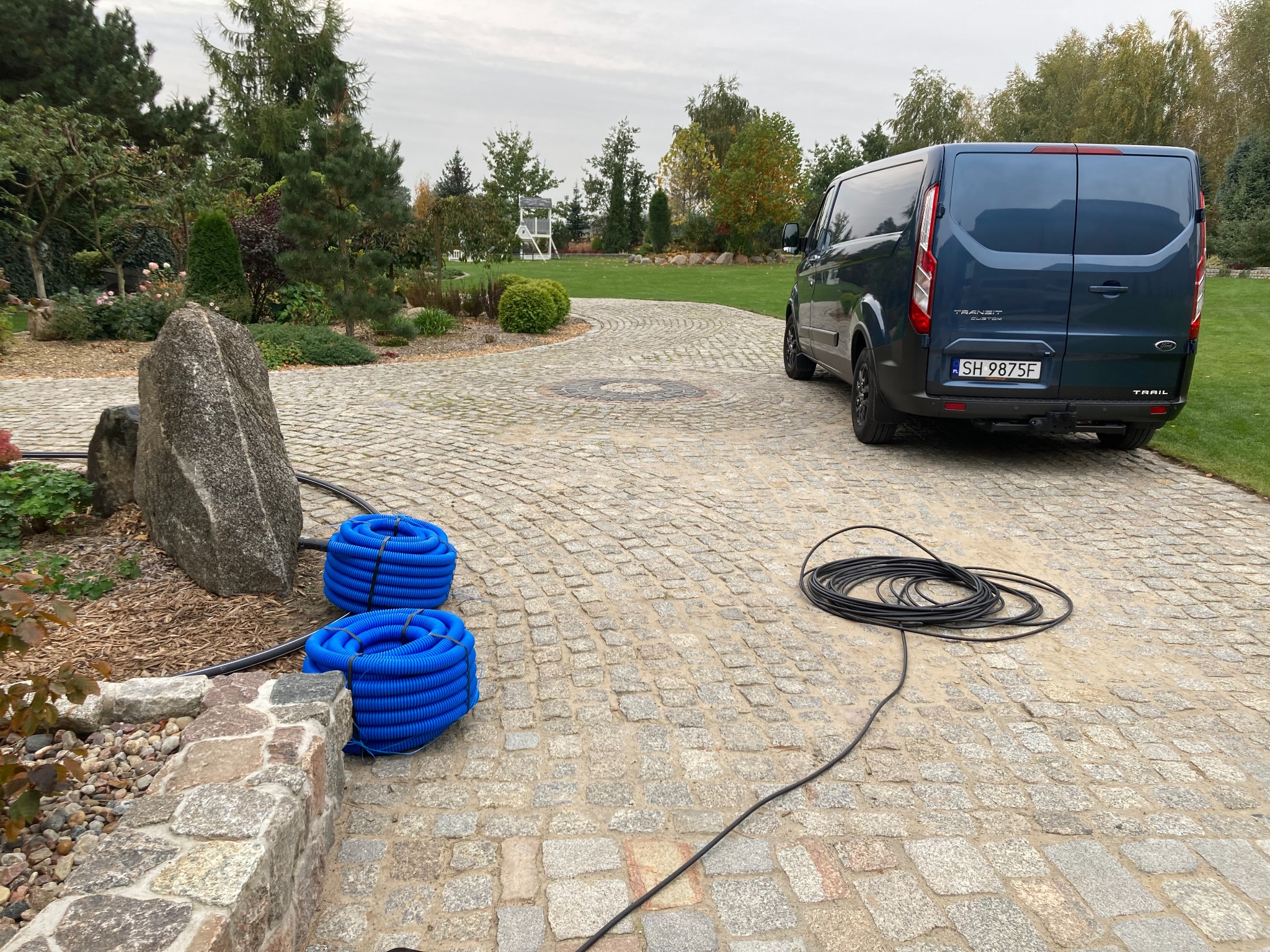 Niebieski bus Ford Transit Custom Trail zaparkowany na brukowanej posesji, obok zwinięte niebieskie rury karbowane i czarny kabel, ogród w tle.