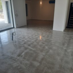 FachMont - Wnętrze w stanie deweloperskim, widoczna wylewka anhydrytowa na podłodze, przygotowana do dalszych prac wykończeniowych, widoczne ślady po szlifowaniu, w tle drabina i otwarte przestrzenie