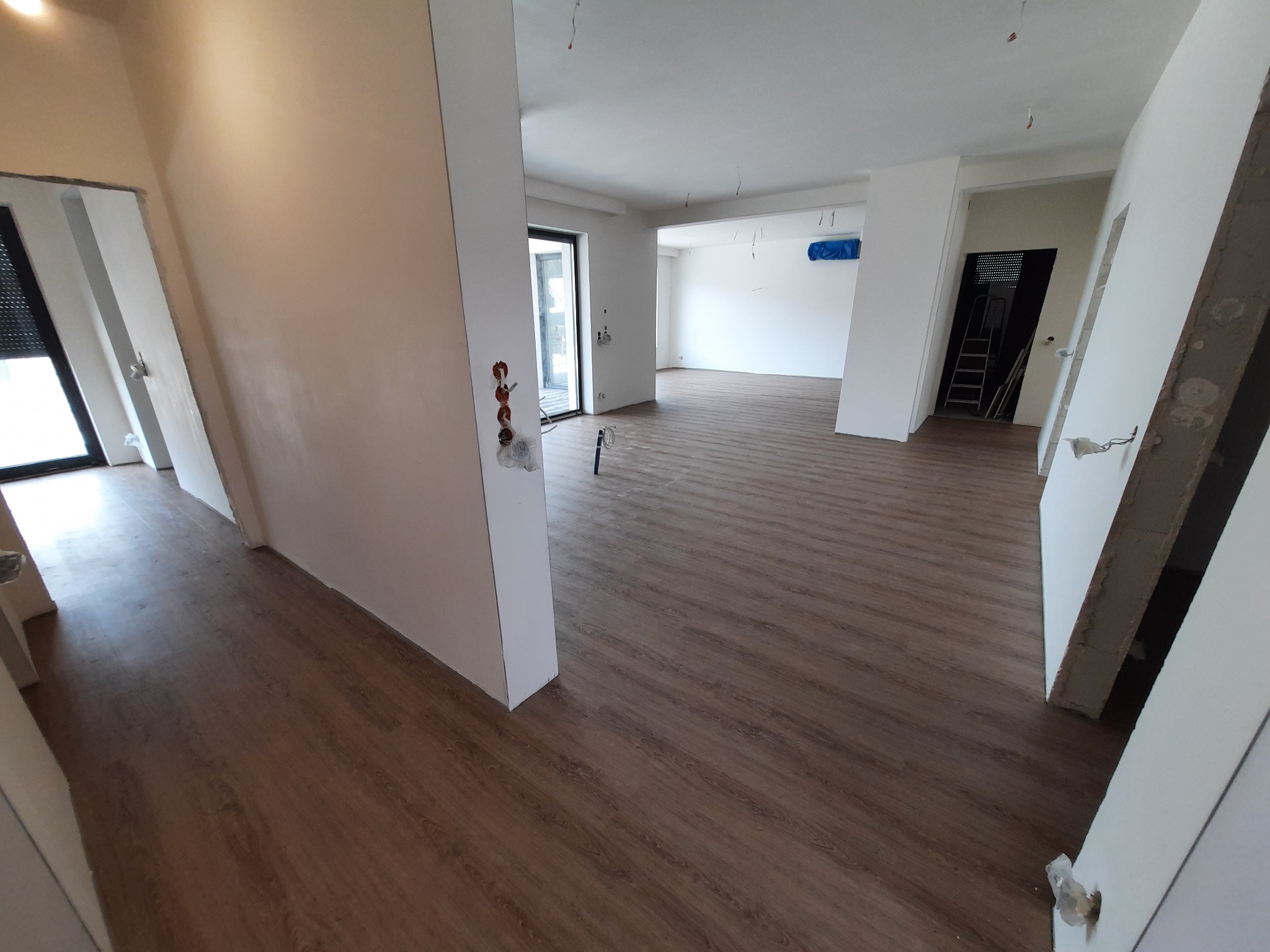 Przestronne wnętrze w trakcie wykańczania, z brązowymi panelami podłogowymi ułożonymi w jodełkę, świeżo pomalowanymi ścianami na biało i widocznymi gniazdkami elektrycznymi bez osprzętu.