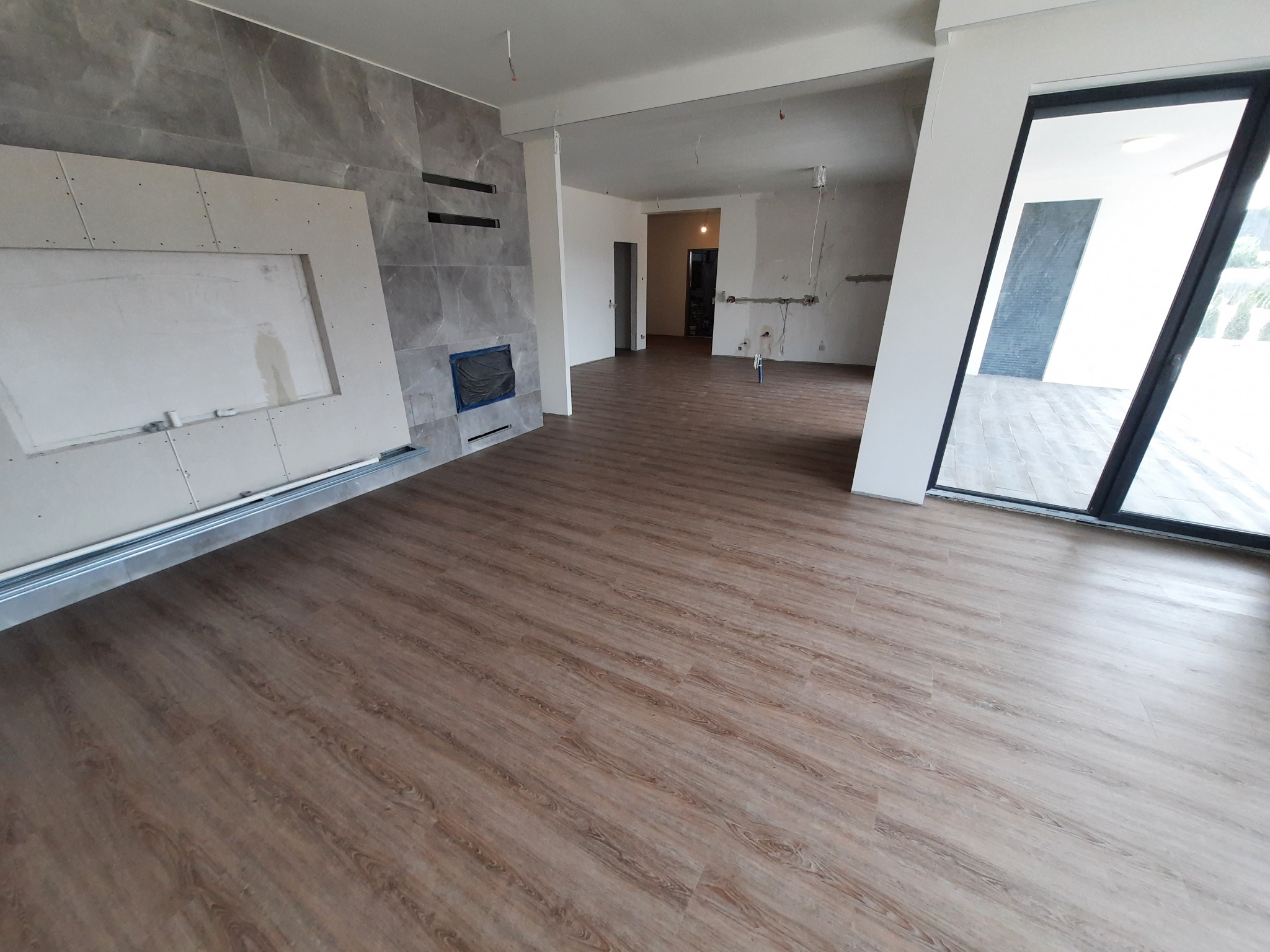 Wnętrze w trakcie wykańczania: ułożone panele podłogowe w odcieniach brązu, ściana z szarą okładziną imitującą kamień i zamontowana płyta kartonowo-gipsowa z wyciętym otworem, widoczne instalacje...