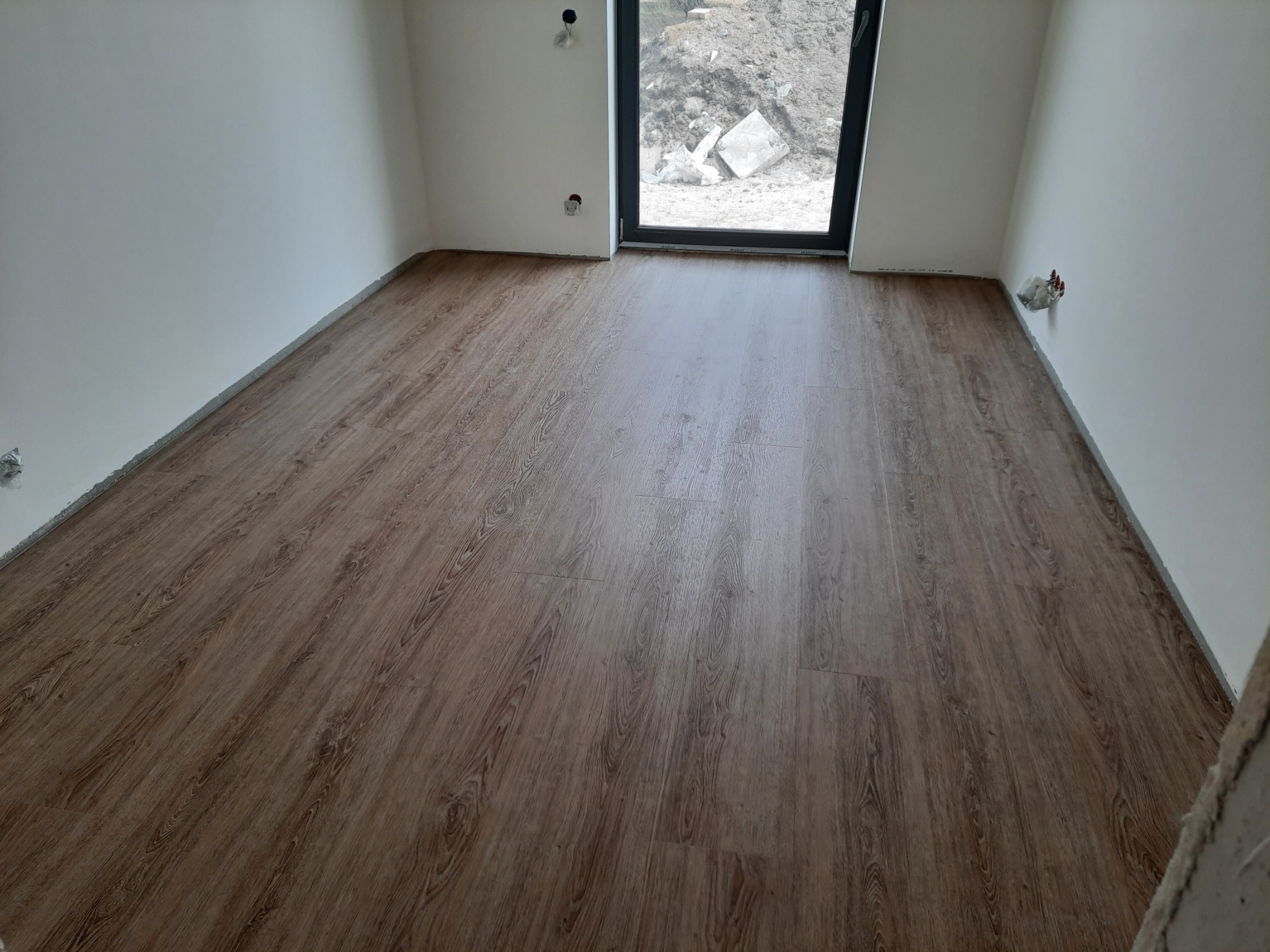 Ułożona podłoga z paneli w pokoju z oknem, widoczne niedokończone wykończenia ścian i gniazdka elektryczne.