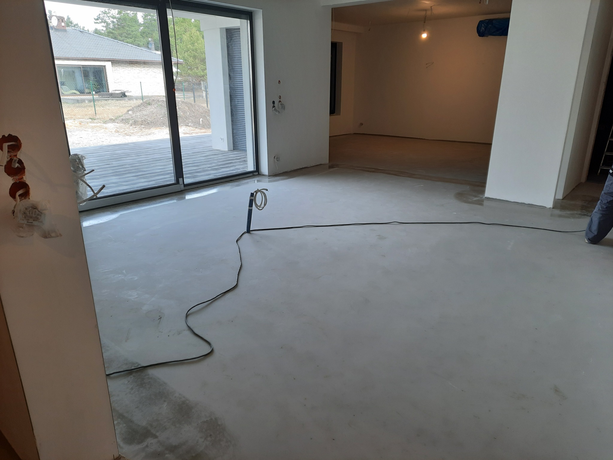 Wnętrze w trakcie prac wykończeniowych, wylewka podłogowa, widoczne kable i instalacje elektryczne, duże okno przesuwne z widokiem na taras i ogród.