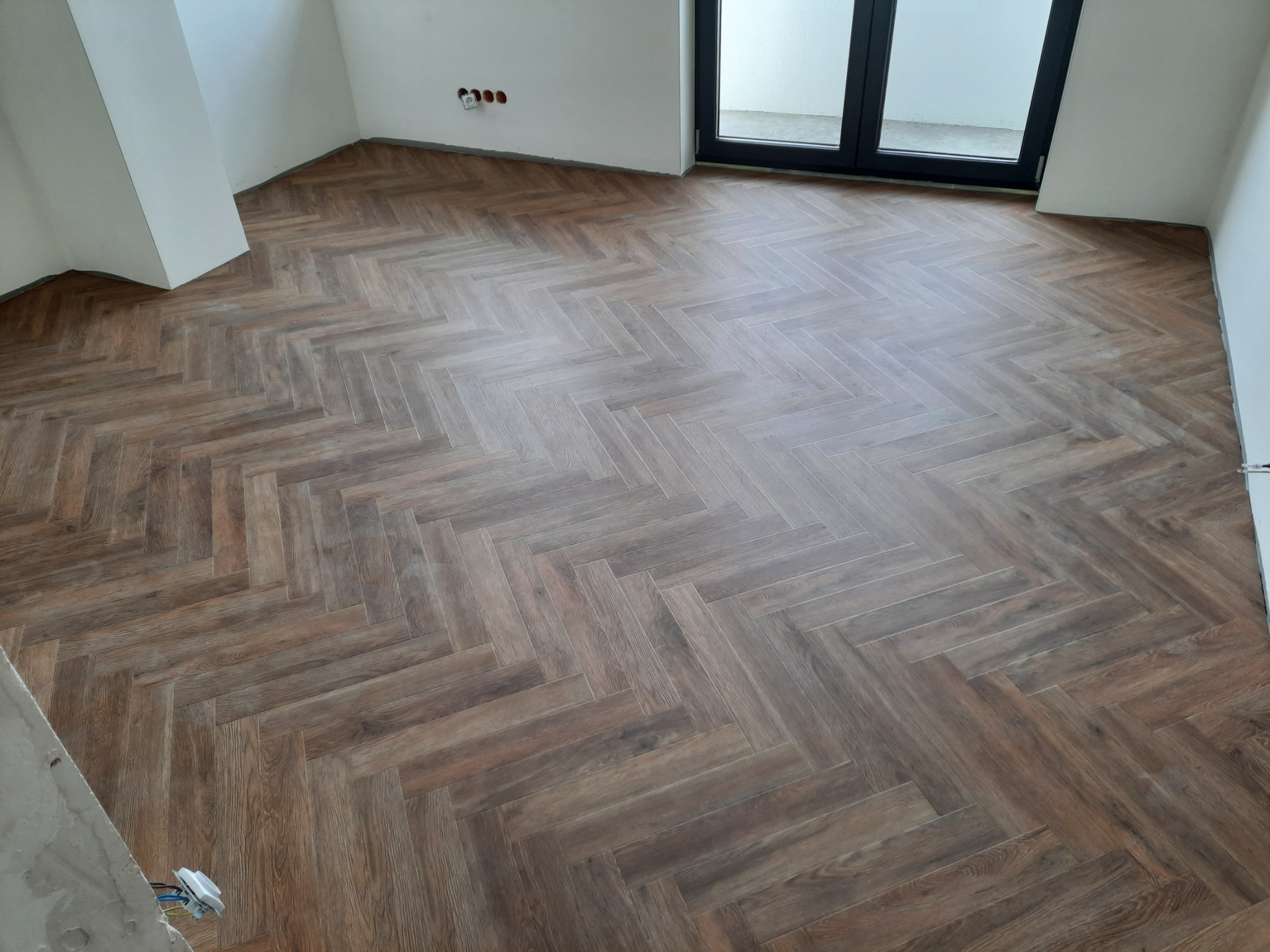 Ułożona podłoga z paneli imitujących drewno w jodełkę francuską, widoczne białe ściany w trakcie wykończenia i okno balkonowe.