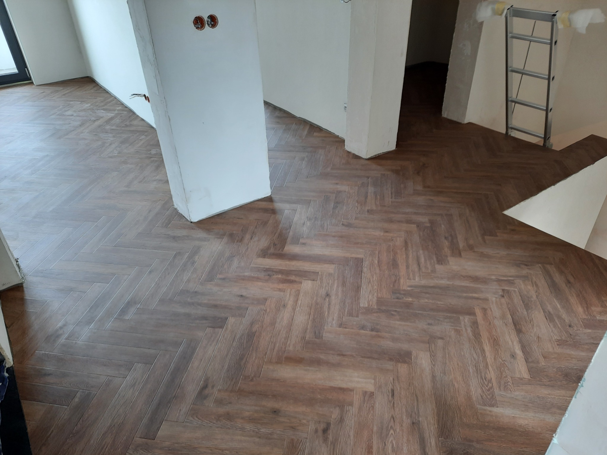 Ułożona podłoga z paneli w jodełkę francuską, widoczne niedokończone ściany i drabina w tle.