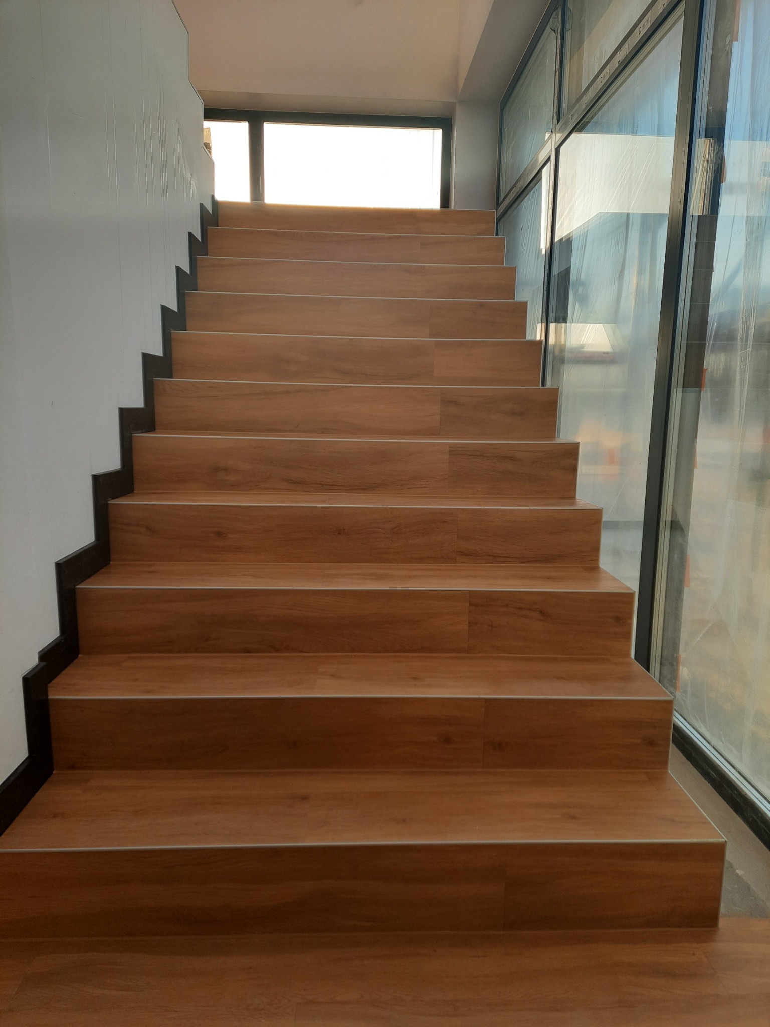 Schody wyłożone panelami drewnopodobnymi z czarnymi krawędziami, widok z dołu, z oknem na górze i szklaną ścianą po prawej stronie.