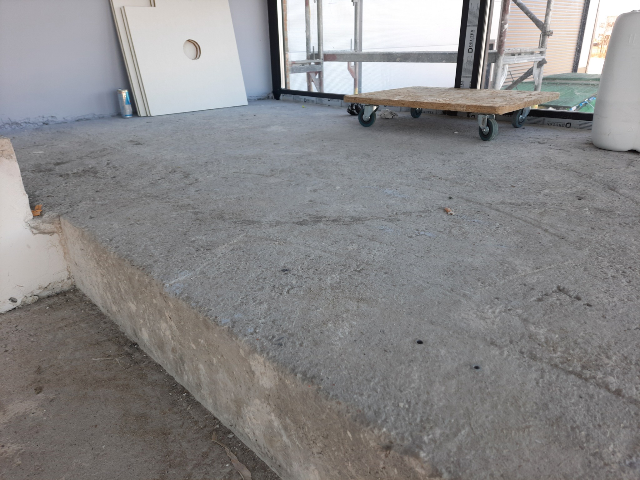 Surowy betonowy podkład podłogowy w trakcie prac wykończeniowych, widoczne płyty drzwiowe i wózek transportowy.