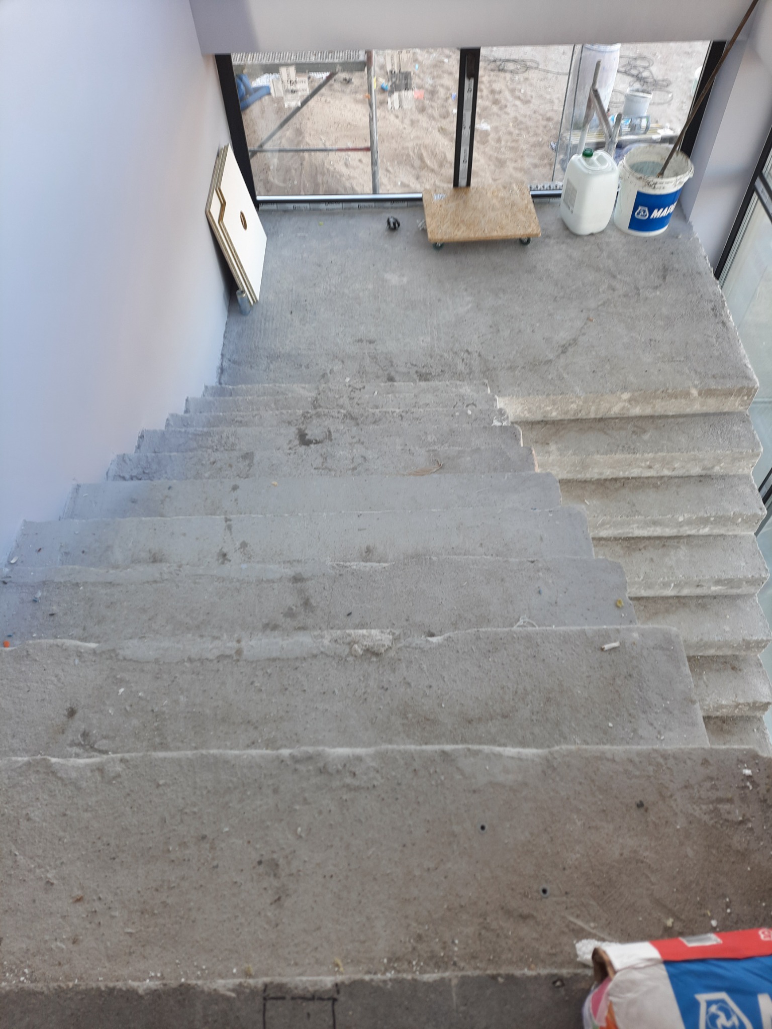 Surowe betonowe schody w trakcie budowy lub remontu, widoczne materiały budowlane i narzędzia, w tle okno z widokiem na zewnątrz.